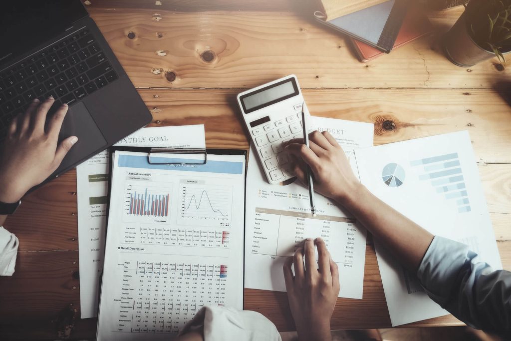 Desk scattered with graphs, a calculator, and a laptop with two people's hands in view.